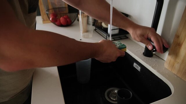 Male hands washing and rinsing a reusable shaker bottle with a sponge in a black kitchen sink