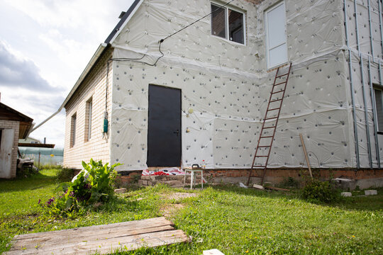 unfinished residential house facade covered with exterior insulation membrane and fasteners, with ladder and entrance door showing ongoing energy retrofit and renovation