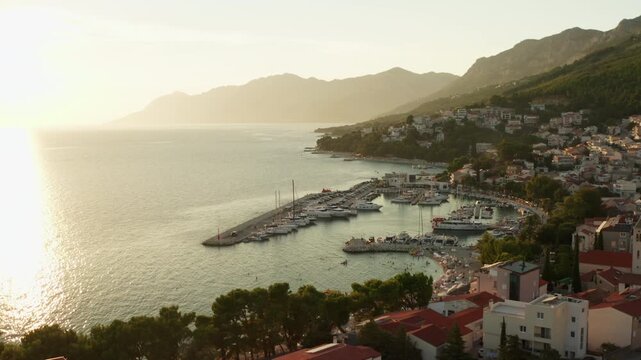 Yachts and sailboats moor in a resort town marina at sunset. Travelers enjoy summer vacation in Baska Voda Croatia. Calm Adriatic sea water reflects golden sunlight along coastal harbor pier.