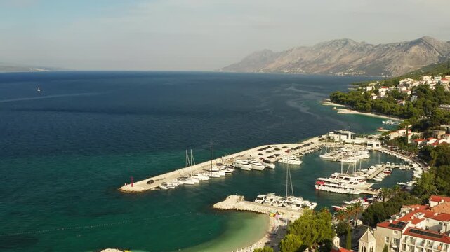 Moored boats and yachts occupy marina berths in Baska Voda resort, Croatia. The nautical port offers seasonal berth rental and secure anchorage for vessels. Calm sea surrounds the harbor pier.