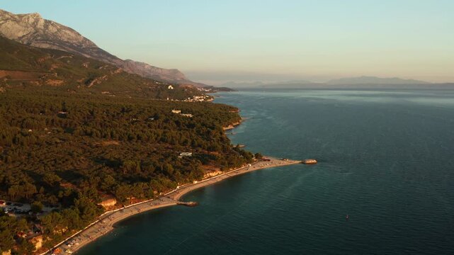 Pine forest surrounds Oseka pebble beach on the Adriatic coast in Baska Voda Croatia. Golden sunset light illuminates the natural cape and clear sea water. Travelers enjoy summer vacation.