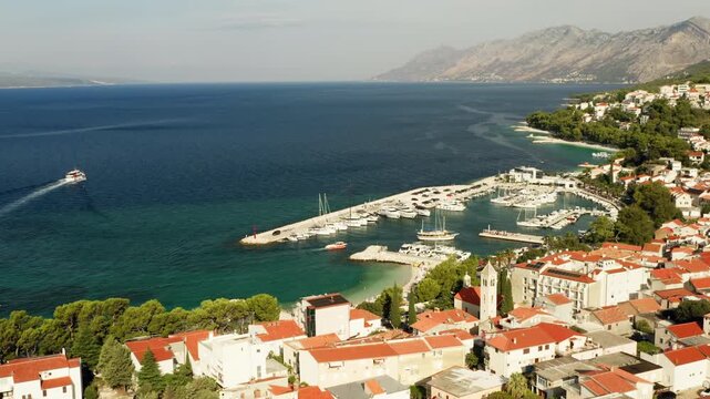 Baska Voda coastal resort town features a sheltered yacht marina harbor in Croatia. Boats moor along the Adriatic Sea waterfront beside dense red roof houses on the Makarska Riviera coast.