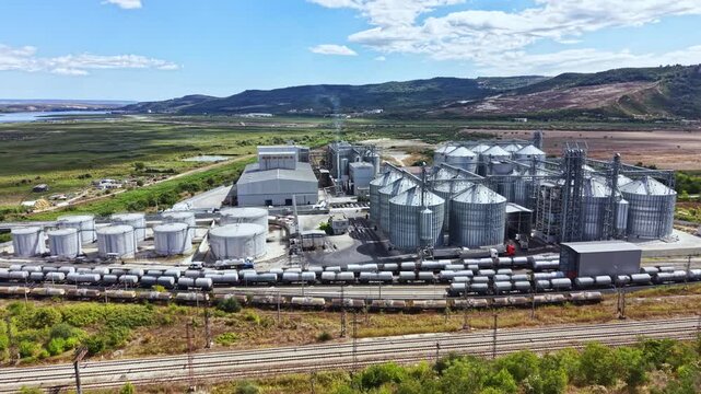 A large industrial site features multiple silos, tanks, and rail cars. It sits next to green fields and mountains under a clear sky during daylight.
