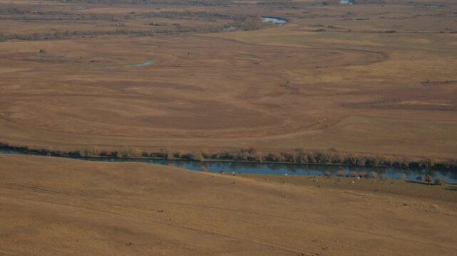 A handheld camera captures a distant view of the winding Bely Iyus river flowing through the expansive, yellowed autumn steppe of Khakassia, Russia. 