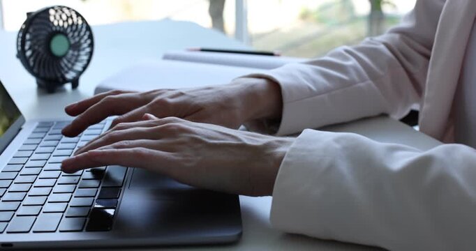 Woman with mini fan working on laptop at white desk in office, closeup