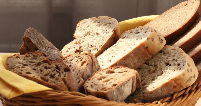Different types of bread in wicker basket on wooden table indoors, closeup. Camera moving in