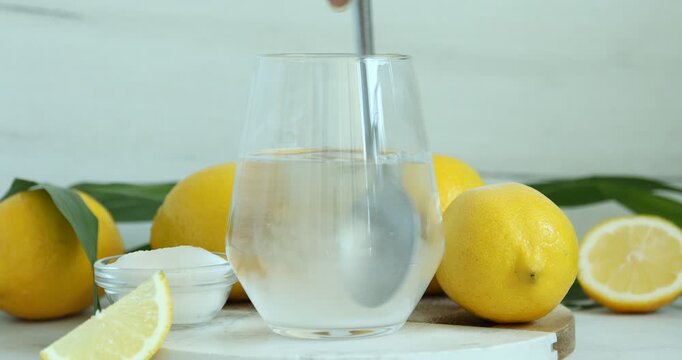 Woman pouring baking soda into glass, stirring it and adding slice of lemon at table, closeup