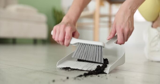 Woman sweeping soil with broom and dustpan on floor indoors, closeup