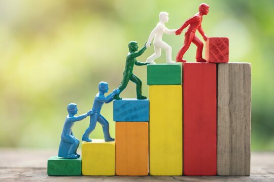 Plastic figures helping each other climb up colorful wooden blocks as a team