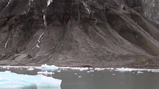 A blue iceberg floats on cold water beneath rugged mountain slopes. Additional visible detail includes foreground detail, with natural light defining the surrounding forms.