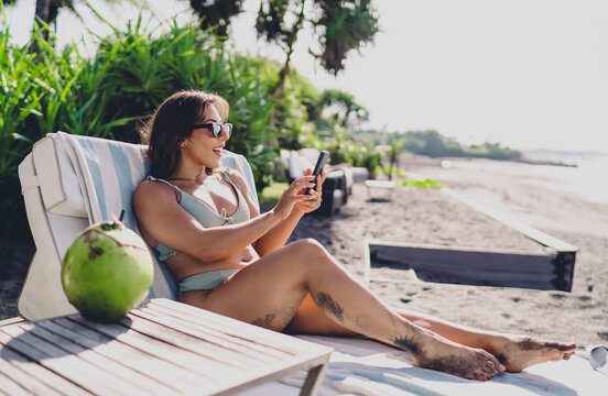 Hispanic female web designer in green swimsuit resting on beach sunbed holding smartphone device, checking mobile application wireframe or website design interface at resort venue space