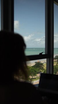Silhueta de pessoa observando a praia atrav&eacute;s de janela de hotel, momento contemplativo e tranquilo durante hospedagem em viagem