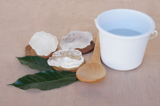 Natural alum crystals displayed with a wooden spoon, green leaves, and a white container on a neutral background, representing traditional household use and natural hygiene.