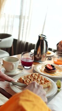 M&atilde;o servindo prato de waffles em mesa farta de caf&eacute; da manh&atilde; com sucos e frutas vermelhas, cena de hospitalidade generosa em viagem