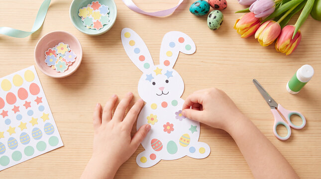 Top view of child hands decorating a paper Easter bunny with colorful stickers, surrounded by craft supplies, painted eggs and spring tulips for a creative seasonal activity