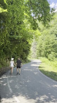 Young couple holding hands, walking away on a winding asphalt mountain road surrounded by lush green forest, vertical video, tripod shot, original unedited 1080 Full HD footage, natural daylight.