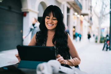 Caucasian female student smiling while operating digital tablet at outdoor terrace table, reviewing e-commerce retail store application discount codes sales and promotional deals in city © BullRun