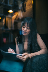 Caucasian female student using stylus pen on digital tablet seen through window glass reflection, listening to digital audiobooks literature streaming media apps and podcast audio in cafe © BullRun