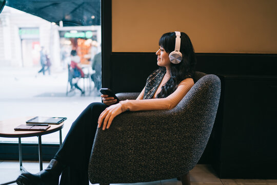 Caucasian female student wearing wireless headphones while sitting in cafe armchair with smartphone, browsing e-commerce retail store discount sales promotional deals and audio offers