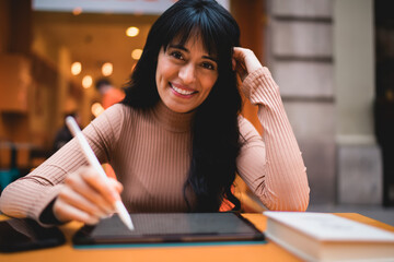 Portrait of smiling Caucasian female freelancer looking at camera while holding stylus pen on tablet screen, creating creative vector artwork deliverables for remote project clients © BullRun