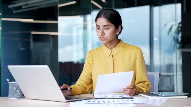 Frustrated businesswoman sitting at desk in office looking at financial report and laptop screen. Upset puzzled female financier checking document with bad indicators disappointed with poor results.