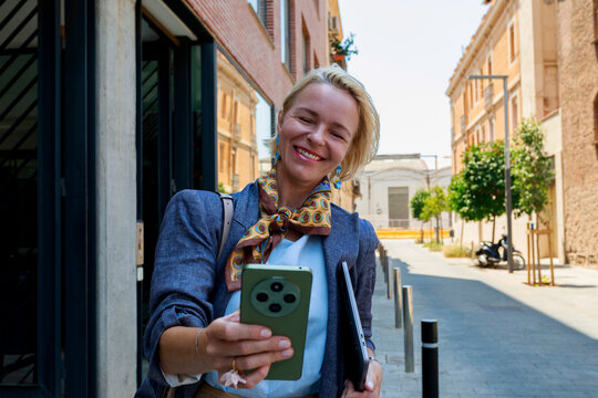 Woman Smiles While Holding Phone on Street in Urban Area