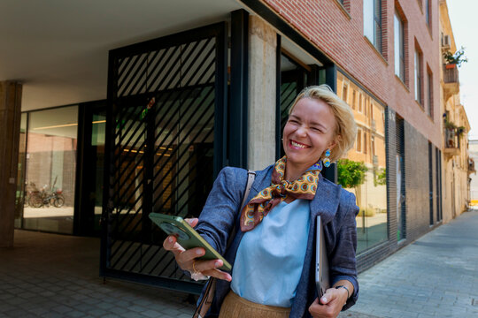 Woman Smiles While Walking on Street Holding Phone and Files
