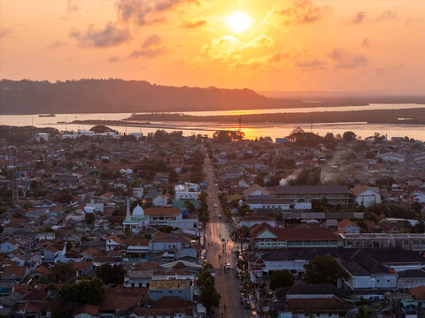 Aerial view of the urban landscape of Cilacap with dense rooftops, a mosque, and a road leading towards the coastal waters under a golden sunset in Cilacap, Central Java, Indonesia.