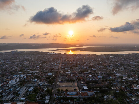 Aerial view of the city of Cilacap at sunset, with the river reflecting the golden light, dense urban rooftops, and clouds in the sky Cilacap, Central Java, Indonesia.