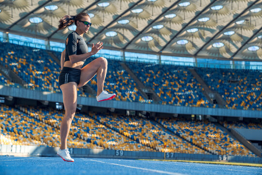 Young woman runner athlete stretching legs before run during sunny morning on stadium track, female sportswoman warming up for training, fitness workout, active lifestyle and race preparation concept