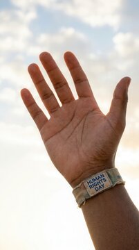 Human rights day bracelet worn on a raised hand against a cloudy sky background.
