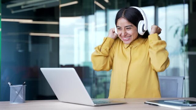Excited businesswoman in wireless headphones sitting at desk in modern office watching sports match or auction on laptop. Happy female cheering, celebrating victory, success, or online achievement.