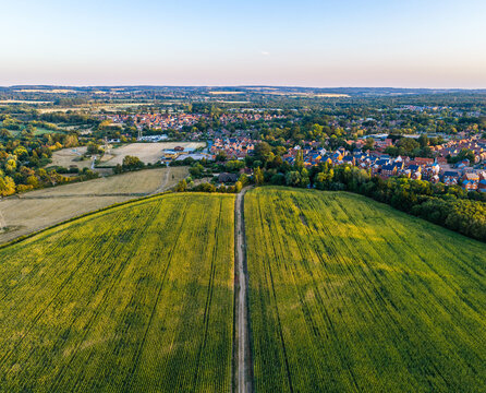 Aerial view of a lush green agricultural field with a central dirt path leading towards a residential village surrounded by trees and rolling hills in Hook, England, United Kingdom.
