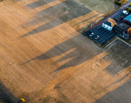 Aerial view of a field with long shadows cast during a solar eclipse, adjacent to a building with a parking lot and a rugby post in Hook, England, United Kingdom.