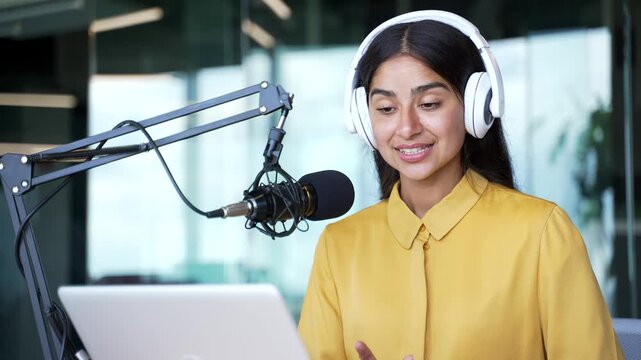 Close-up of young businesswoman in headphones speaking into microphone while using laptop in modern office. Confident female professional recording podcast, hosting online webinar or virtual interview