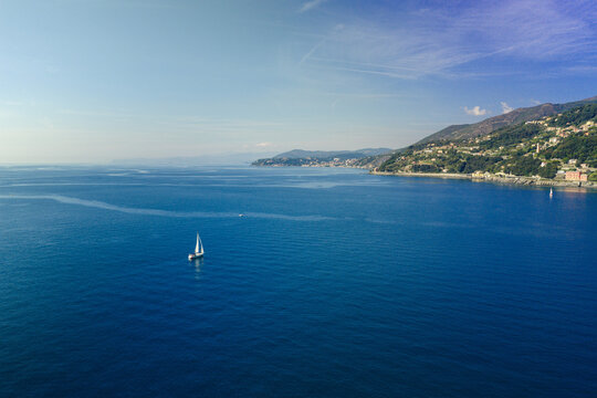 Aerial view of a lone sailboat gliding across the deep blue waters of the Ligurian Sea near the rugged coastline of the Italian Riviera, Genova, Liguria, Italy.