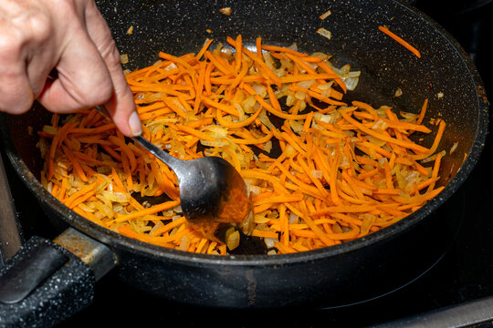 Hand stirring sauteed carrots and onions in a black frying pan on a stovetop, with vibrant orange and yellow colors visible in the cooking process