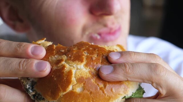 Man eats sandwich with both hands, lifting it to his mouth in repeated bites. Close framing emphasizes fresh bread, visible fillings, and steady concentration while he enjoys lunch.