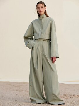 Young woman standing in a desert-like environment with a plain white wall in the background. she is wearing a long, light green jumpsuit with a high collar and long sleeves.