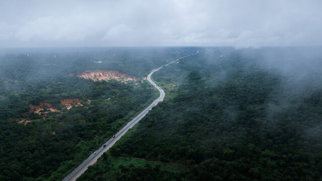 Aerial view of the Owerri - Orlu Road winding through a dense, lush green forest under a cloudy, misty sky in Owerri, Imo, Nigeria.