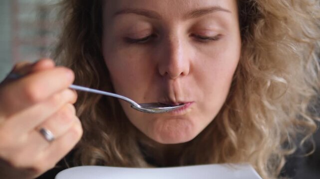 Woman tastes creamy soup with spoon from white bowl, smiling with satisfaction. Hungry diner lifts each spoonful, eats slowly, and enjoys warm comfort food topped with crunchy croutons.