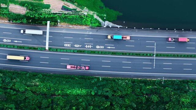 High angle top down aerial view of multi lane highway with freight trucks traveling past green vegetation and body of water.