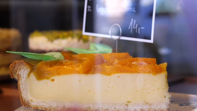 Cream cake slice rests behind bakery glass. Dessert slice sits on display with handwritten price tag, orange topping and green garnish, suggesting fresh pastry counter service and sweet indulgence.