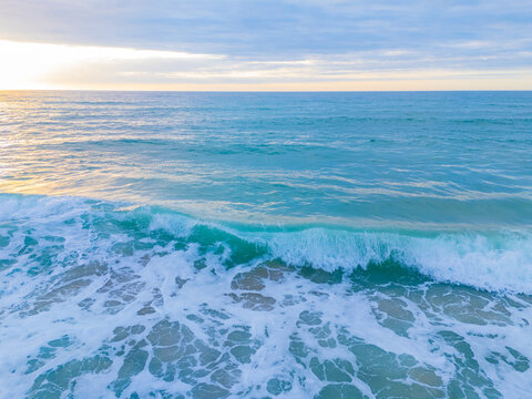 Aerial view of turquoise ocean waves breaking on the sandy shore at sunrise with soft light reflecting on the water in Wrightsville Beach, North Carolina, United States.