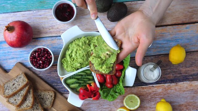 Hands assemble avocado toast on rustic tabletop with knife and bright produce. Avocado spread, bread slices, tomatoes, cucumber, citrus, and pomegranate create fresh, upbeat breakfast mood.