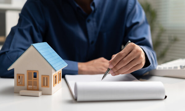 Real estate businessman reviews property documents beside a house model, representing home purchase, mortgage, contract, property investment, financing, ownership, and housing agreement.