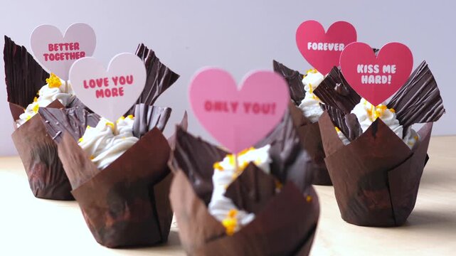 Valentine style cupcakes stand ready on clean tabletop, each topped with hearts reading love notes. Dessert arrangement looks festive and tender, with rich chocolate wrappers and cream frosting.