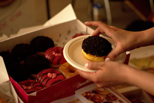 Person holding a chocolate sprinkled donut on a small plate over a box of assorted colorful donuts.