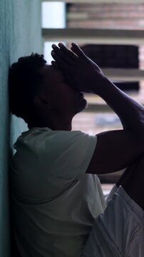 African American man sitting on ground leaning against wall with hand on forehead in deep stress