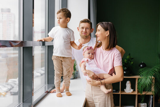 Happy Family with Two Little Children Standing by Window at Home